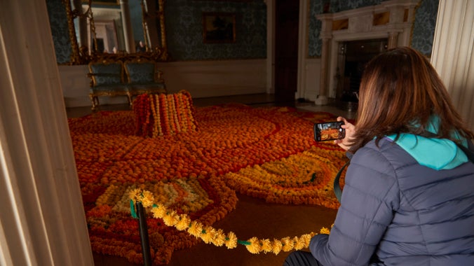 A display of colourful rangoli artwork is displayed flowing across the floor of the historic house room. It is made from flowers and other materials, mostly shades of orange or yellow